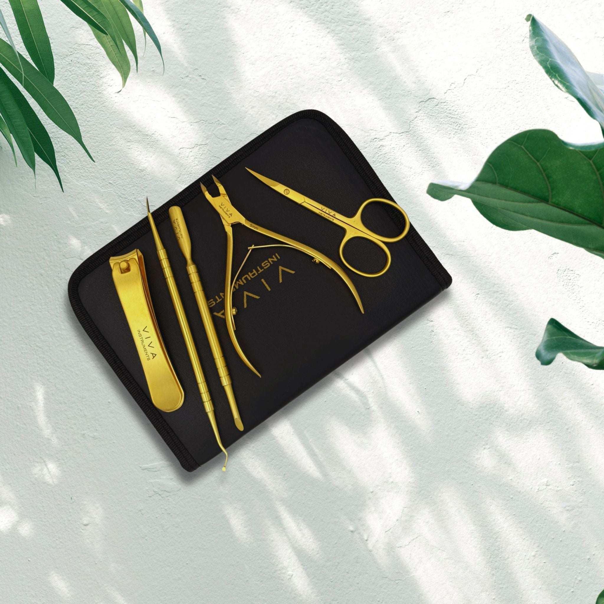 Set of gold scissors and tools on a black case with 'Vita' branding, placed on a light surface with green leaves. viva instruments