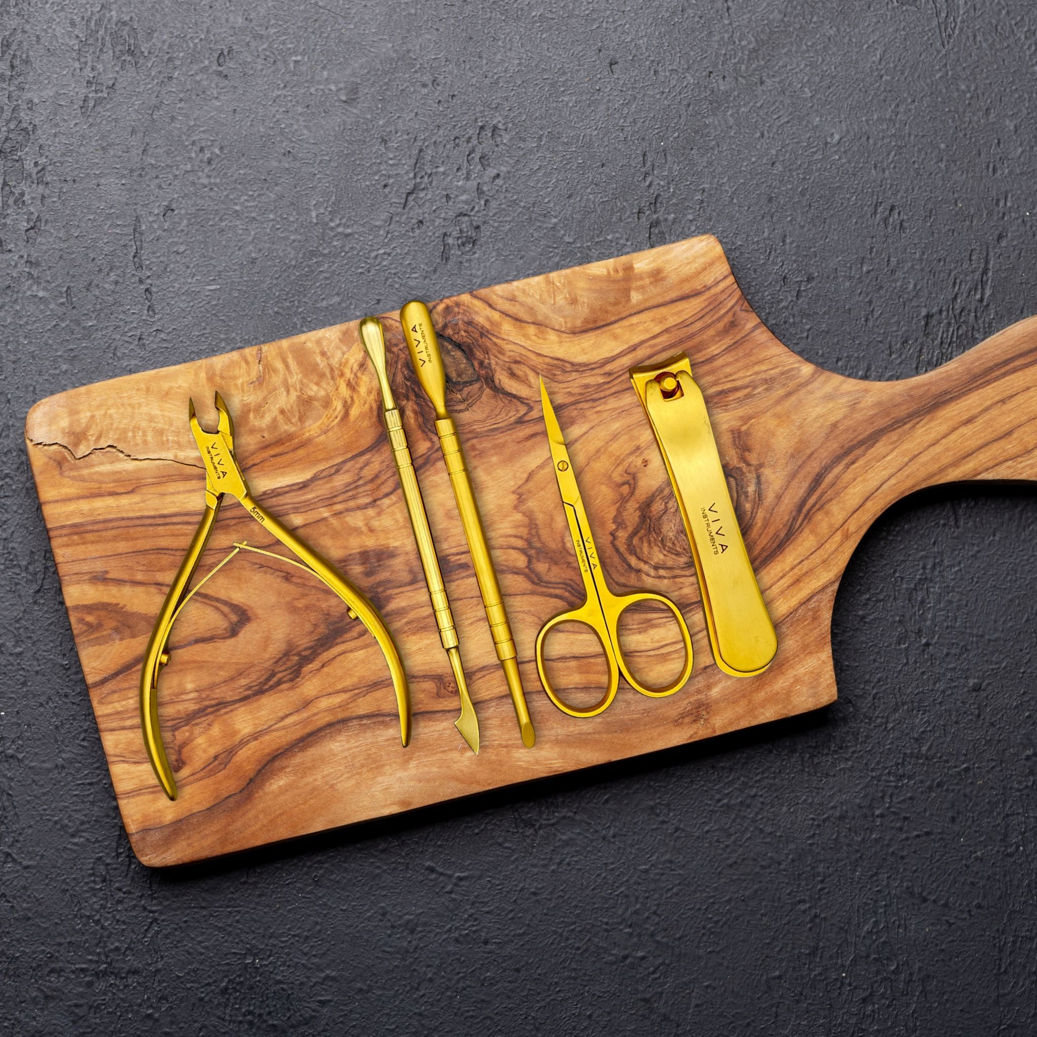 Set of gold scissors, cuticle nippers, nail clippers, & pushers on a wooden cutting board against a dark background - viva instruments